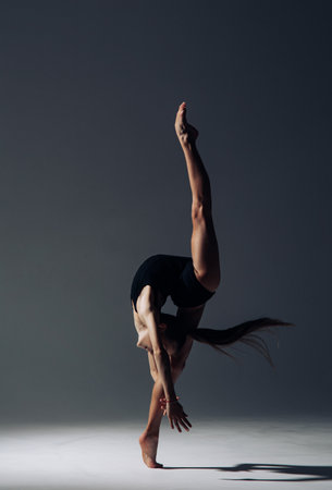 Gymnast child girl in black bodysuit trains lit by light on dark background.の写真素材