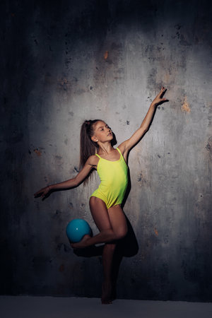 Gymnast child girl lit by light in yellow bodysuit performs gymnastic exercises with ball on dark background.の写真素材