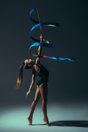 Gymnast child girl lit by light in black bodysuit performs gymnastic exercises with ribbon in her hand on dark background.の写真素材
