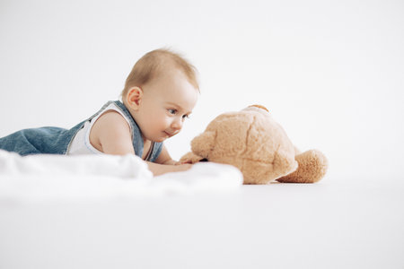 Cute baby boy lies on floor and plays with toy plush bear against white background.の写真素材