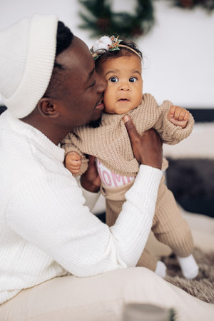 Happy african man holds and kisses his mixed race baby daughter against background of Christmas decorations. Concept of interracial family and unity between different human races.の写真素材