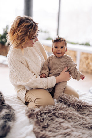 Happy caucasian woman sits on bed and plays with her mixed race baby daughter against background of...