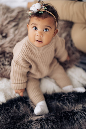 Surprised mixed race baby girl sits on bed with wide-eyed. Baby has wreath with Christmas decoration...