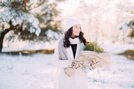 Happy young smiling woman walks in forest among snow covered pine trees in sunny winter day with bouquet in her hands.の写真素材