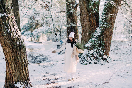 Happy young smiling woman walks in forest among snow covered trees in sunny winter day with bouquet in her hands.の写真素材