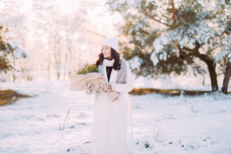 Happy young smiling woman walks in forest among snow covered pine trees in sunny winter day with bouquet in her hands.の写真素材