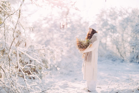 Happy young woman walks in forest among snow covered trees against background of snowflakes with bouquet from pine branches in her hands.の写真素材