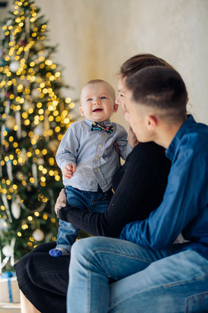 Happy parents play with their baby son against background of Christmas tree with decorations and glowing garlands. Image with bokeh effect.の写真素材