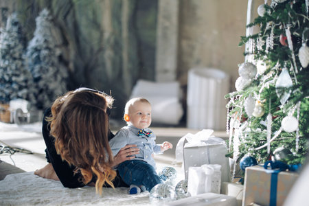 Happy mother plays with her baby son near Christmas tree with decorations and gift boxes.