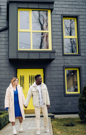 Young interracial couple holds each other hand and walks on street against background of houses. Concept of love relationships and unity between different human races.の写真素材