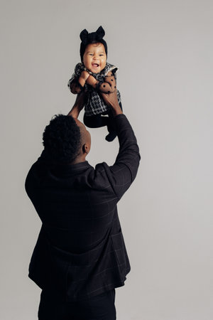 Young african man plays with his baby daughter from interracial marriage against gray background. Baby laughs cheerfully. Concept of interracial family and unity between different human races. back view.の写真素材