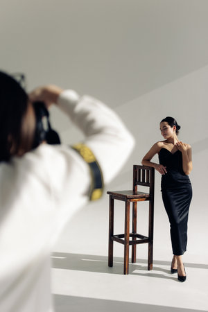 Photographer shoots young woman posing and relaxing with closed eyes in studio near chair in long black dress and high heel shoes against sunlit background.の写真素材