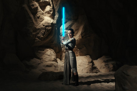 Kyiv, Ukraine - April 14, 2023: Cosplayer girl portrays character Rey Skywalker from the Star Wars universe with blue blade lightsaber in her hand among sandy canyon.のeditorial素材