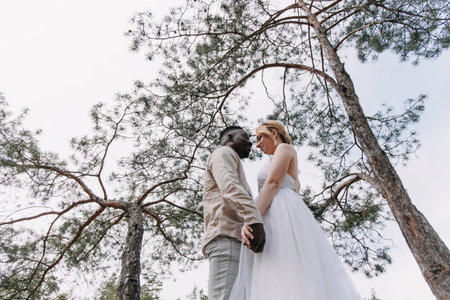 Happy interracial couple newlyweds stands and holds each other hands against background of pine trees and sky. bottom view. Concept of love relationships and unity between different human races.の写真素材