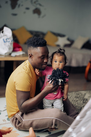 Young african man plays with his baby daughter from interracial marriage in room among toys on floor. Concept of interracial family.の写真素材