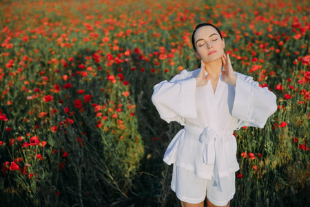 Young woman in white clothes walking among flowering poppies and enjoying meadow with closed eyes at sunset. Image with copy space.の写真素材