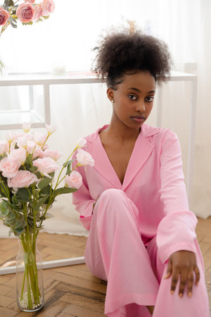 Young african pensive woman in pink pajamas sitting in floor near flower bouquet against background of window.の写真素材