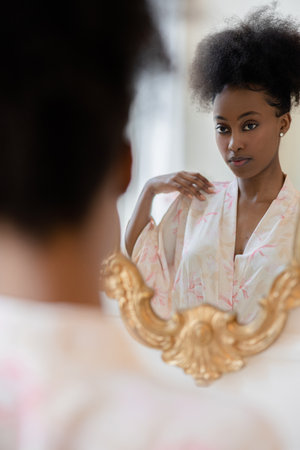 Young african woman in pajamas standing against mirror and looking at her reflection in it against background of home interior. closeup. back view.の写真素材