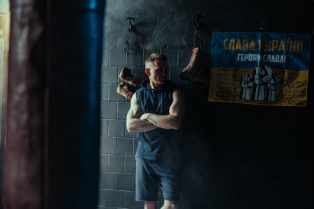 Tired boxer resting in dark gym after exercising against background of Ukrainian flag with...