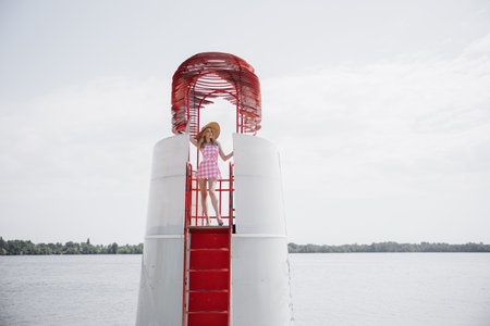 Young blonde girl look like a  doll in pink checkered mini dress and wide brim hat standing on lifeguard beach tower against water and sky background.の写真素材