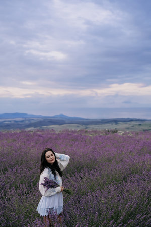 Young brunette woman walking with bouquet of lavender in her hands against background of blooming lavender field and sky.の写真素材