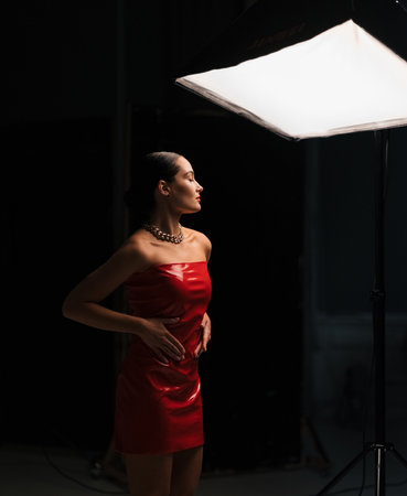 Young pretty brunette woman poses in stylish red latex dress under spotlight in studio against black background.の写真素材