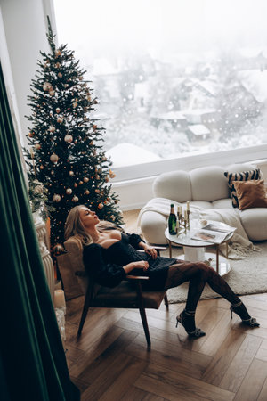 Young pensive blonde woman sitting in armchair in stylish black mini dress with decollete near decorated Christmas tree and table with champagne bottle against background of window.の写真素材