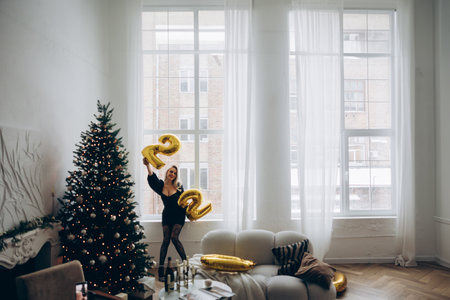 Young smiling blonde woman standing near Christmas tree in stylish black mini dress with decollete...