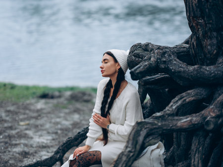 Young dreamy woman with closed eyes and braided hair sitting and resting in forest near trunk tree in white sweater, skirt and hat against background of lake.の写真素材