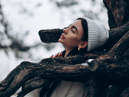 Portrait of young dreamy woman with closed eyes sitting and resting in forest near trunk tree in white sweater and hat. Closeup.の写真素材