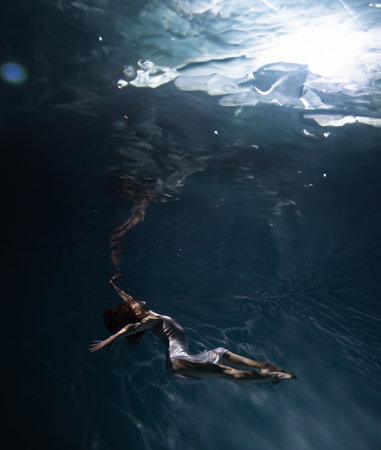 Underwater shoot of beautiful ballerina in white dress swimming and dancing in water through...