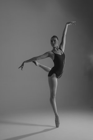 Young ballerina in bodysuit and pointe shoes performing Arabesque pose against gray background. Graceful beautiful ballerina. Ballet art, grace, flexibility, motion. Black and white image.の写真素材