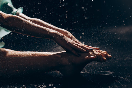 Close-up view to legs and arms of ballerina girl sitting under falling water drops on wet floor against dark background. Graceful beautiful ballerina. Ballet art, grace, flexibility, motion.の写真素材