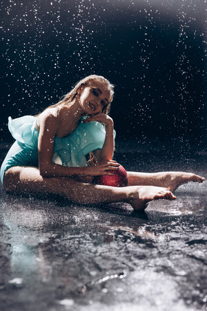 Ballerina teenager girl in green bodysuit dances with ball against falling water drops and light of spotlight on dark background. Graceful beautiful ballerina. Ballet art, grace, flexibility, motion.の写真素材