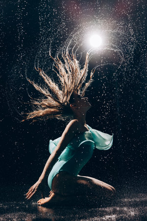 Ballerina teenager girl in green bodysuit dancing under falling water drops and shakes her wet hair against spotlight on dark background. Graceful ballerina. Ballet art, grace, flexibility.の写真素材