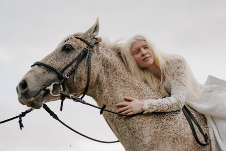 Closeup and bottom view of young albino woman rides horseback on gray horse in white dress against...