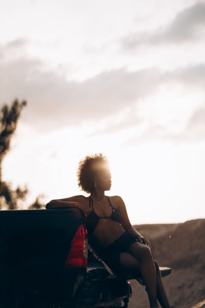 Young African woman sitting on open pickup truck bed against background of sandy canyon at sunset.の写真素材
