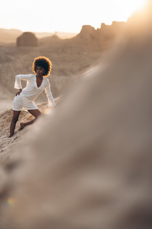 Pretty young African woman posing in white dress against background of sandy canyon at sunset. Backlit.の写真素材
