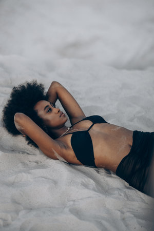 Young sad african woman posing in black bra and skirt lying down on sand. Top view.の写真素材