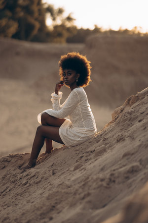 Pretty young African woman sitting in white dress in sandy canyon at sunset. Backlit.の写真素材