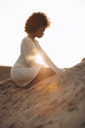 Young African woman sitting in white dress in sandy canyon against background of sunset sky. Closeup.の写真素材