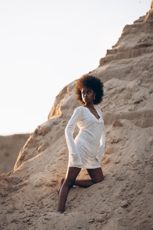 Pretty young African woman posing in white dress against background of sandy canyon at sunset.の写真素材