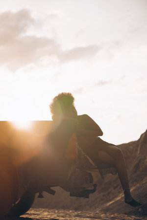 Young woman sitting on open pickup truck bed against background of sandy canyon at sunset.の写真素材