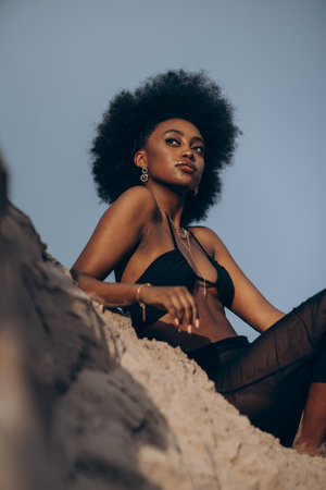 Pretty young African woman posing in black bra and skirt against background of sand and blue sky. Closeup. Bottom view.の写真素材