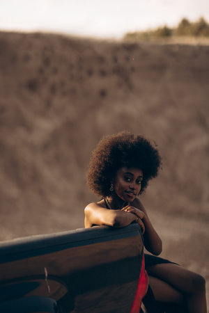 Pretty young African woman sitting on open pickup truck bed in black bra and skirt against background of sandy canyon at sunset.の写真素材