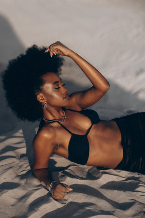 Pretty young African woman posing with closed eyes in black bra and skirt lying down on sand on beach. Top view.の写真素材