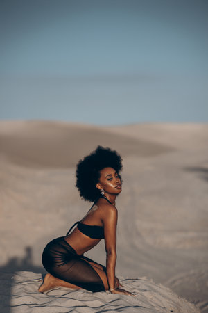 Pretty young African woman posing in black bra and skirt against background of sandy desert and blue sky. Closeup.の写真素材