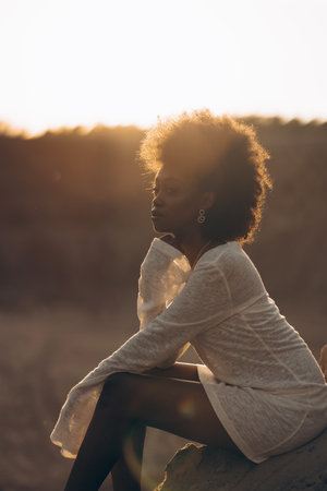 Pretty young African woman sitting in white dress against background of sunset sky. Backlit. Closeup.の写真素材