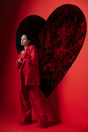 Beautiful girl in red pants and jacket posing on red background decorated by heart shape with red roses. Holiday party, birthday.の写真素材