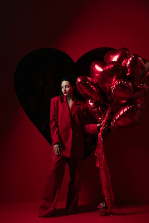 Beautiful girl in red pants and jacket posing against big black heart on red background with heart shape air balloons in her hands. Valentines day. Holiday party, birthday.の写真素材